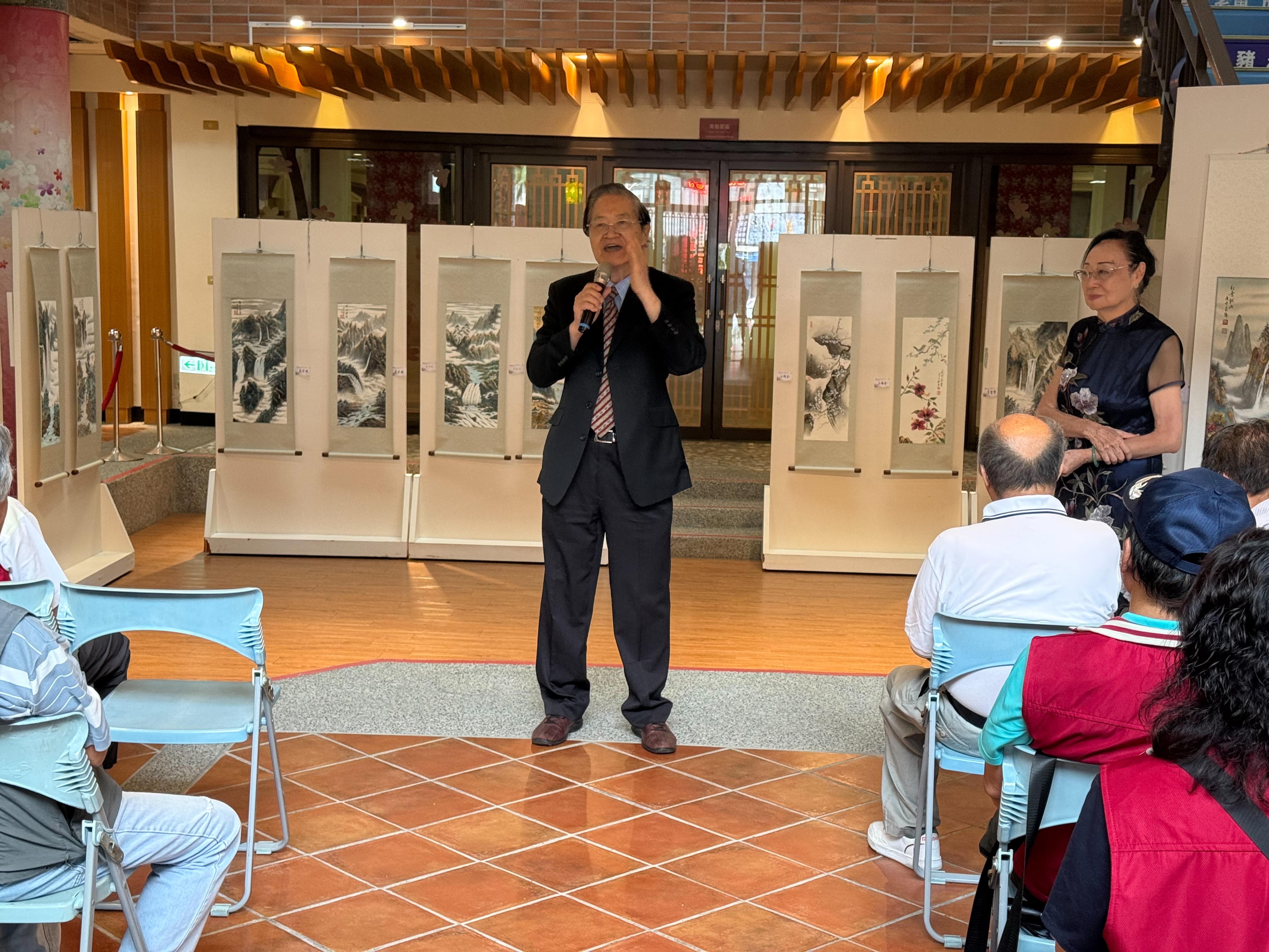 策展人張志強教授於「墨舞丹青」張志強師生聯展開幕儀式致詞，後方陳列多幅展出作品。（圖／臺南市政府客家事務委員會提供）